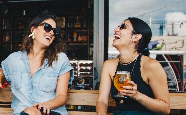 Två kvinnor i solglasögon dricker öl och skrattar. Foto.