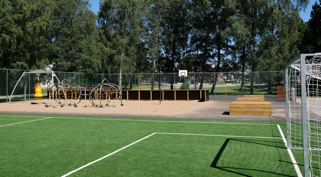 Bilden är en översiktsbild över De Lavals aktivitetsyta. I förgrunden syns en fotbollsplan. I bakgrunden, till vänster, syns två runda ställningar för klättring. I bakgrunden i mitten av bilden syns en basketkorg. Bilden är tagen på sommaren och solen lyser.  