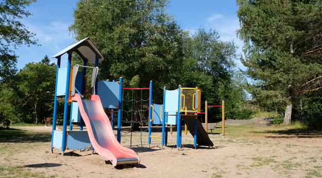 Bilden är ett fotografi över en lekplats. Till vänster i bild syns en lekställning med en rutschkana. Kanan är röd och huset blått. Till häger i bild syns en till lekställning. Denna ställning har en klättervägg och en liten utkiksplats. 
