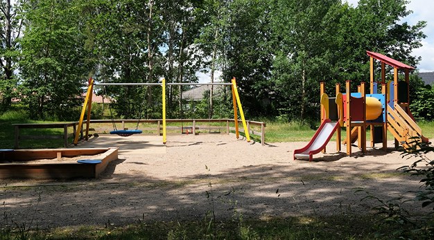 Bilden är en översiktsbild över Karolinerparkens lekplats. Från vänster i bild är det en sandlåda, en gul gungställning och en stor lekställning med två rutschkanor. Marken under är sand. 