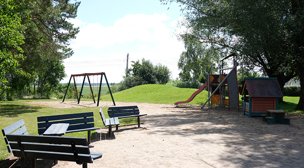 Fotografiet är en översikts bild över Lundhagens lekplats. Till vänster i förgrunden syns fem blå parkbänkar. Varav tre står som i en halvcirkel. Bakom parkbänkarna syns en röd gungställning. Till höger i bild står en lekställning och en liten lekstuga. Bakom lekställningen är en lite mindre gräsklädd backe. Fotografiet är taget på sommaren och solen lyser.  