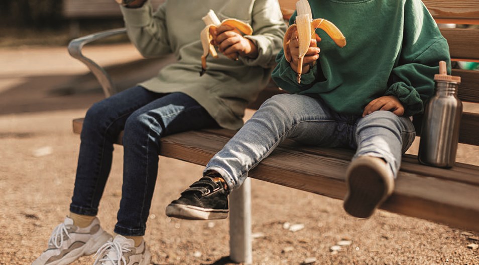 Bild på fyra barnsben som sitter på en parkbänk och äter banan. Bilden är beskuren vid axlarna på barnen.