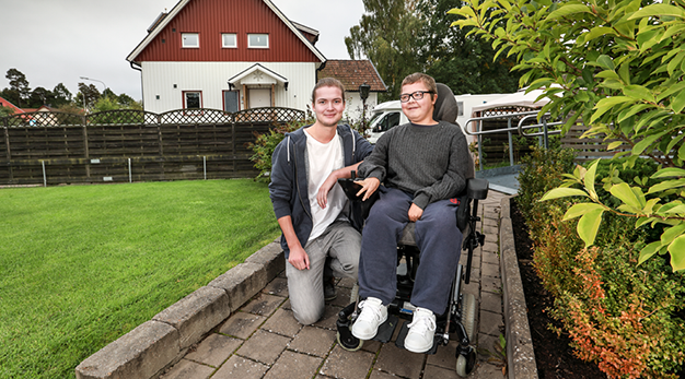 Bild på en personlig assistent tillsammans med en yngre kille i rullstol på uppfarten till ett hus.