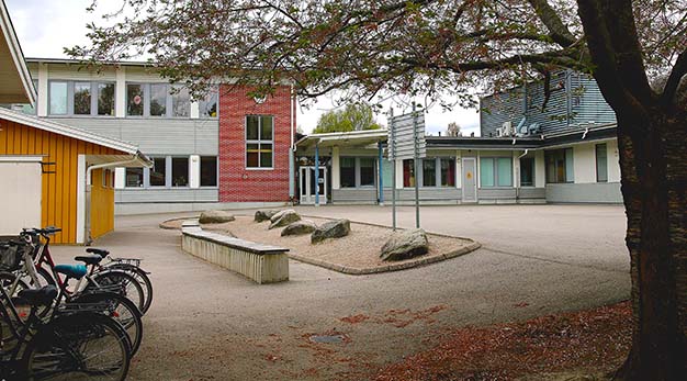 Bilden föreställer en entré till Hjortmosseskolan. I förgrunden syns en skolgård och till höger i bild står en stort träd. På marken nedanför ligger en stor hög med löv. Fotografiet är taget på hösten. Skolans fasad i höger i bild är ljusgrå och vit. Denna del sitter ihop med en större byggnad som är i tegelsten med gråa träpartier. På skolagården finns också en liten gul byggnad.