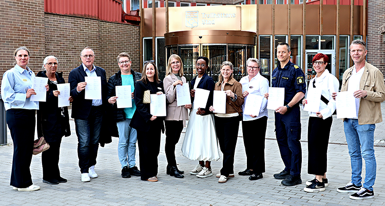 grupp människor står och tittar in i kameran. dE håller varsitt papper framför sig. 