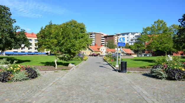 Bilden föreställer ingången till Betty Backs park. Bilden är tagen från gångstråket. Underlaget är tegelsten. På båda sidorna av gångstigen finns stora öppna ytor med gräsmatta. I förgrunden står två stora krukor med blommor av olika slag. Gula, svarta, rosa och gröna. Bilden är tagen på sommaren och solen skiner.