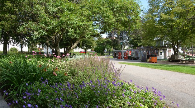 Bilden föreställer en park. Till vänster i bild är det en stor rabatt med flera olika blommor och buskar i vitt, grönt, lila och rosa. Bakom rabatten finns ett träd med en stor krona. Till höger i bild löper en gångväg som leder fram till macken-lekplatsen. I bild syns Roy och Rogers verkstad. Fotografiet är taget på sommaren och solen lyser. 