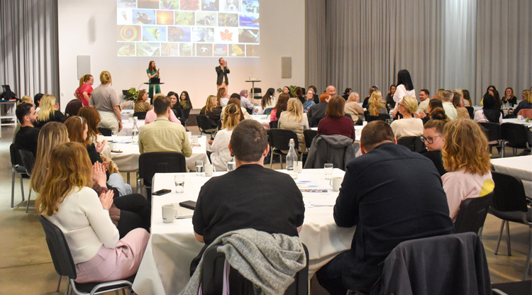 En stor grupp människor sitter vid dukade bord i en hörsal och lyssnar på två personer som talar på en scen med en projektorbild i bakgrunden.