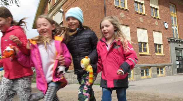 Fotografiet föreställer fyra flickor som springer mot kameran. I bakgrunden syns fasaden på en skolbyggnad. Barnen ler stort. 