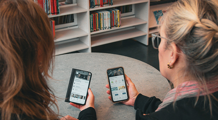 Två tjejer tittar på var sin telefon inne i ett bibliotek. Foto.