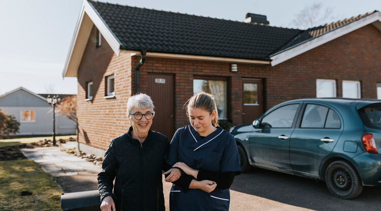En kvinna får stöd av en undersköterska ut på promenad. Kvinnan ler. Foto.