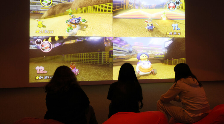 tre ungdomar spelar mario cart på storbildskärm