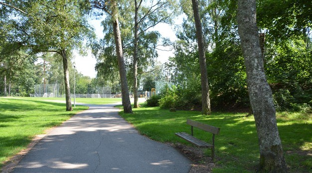 Bilden är ett fotografi på en asfalterad gång- och cykelväg. Längst sidorna är det gräsmatta och grönska i form av buskar och träd. Till höger lägst cykelvägen står en parkbänk i mörkt trä. Långt i bakgrunden syns en fotbollsplan. 