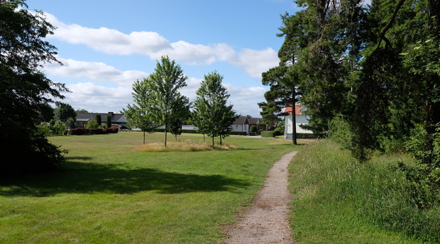 Bilden föreställer en liten grusstig som löper bredvid en park. Till vänster i bild är det en stor öppen yta med tre träd och till höger är det buskar och träd. 