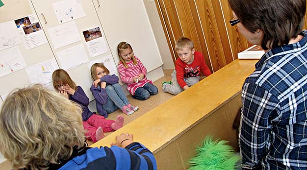 fyra barn sitter och tittar på dockteater som två fröknar spelar.