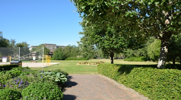 Bilden föreställer Dahllöfs park. Parken är formad som en rektangel och fotografiet är taget från ena kortsidan. I förgrunden till vänster i bild är en rabatt belägen. Det är flera olika typer av blommor och buskar i rabatten, blommorna är lila och rosa och buskarna skiftar i olika typer av grönt. Till höger i bild är en grön välklippt häck och ur denna växer två stora träd. I bakgrunden finns en stor öppen yta med gräs. Det finns även tre bänkar. Till vänster i bakgrunden syns en volleybollplan. 