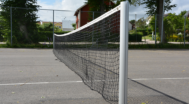 Fotografiet är en närbild över ett tennisnät. Bilden är tagen från sidan så nätets ställning är i fokus. Marken är asfalt och i bakgrunden syns ett högt gallerstaket och en husknut. 