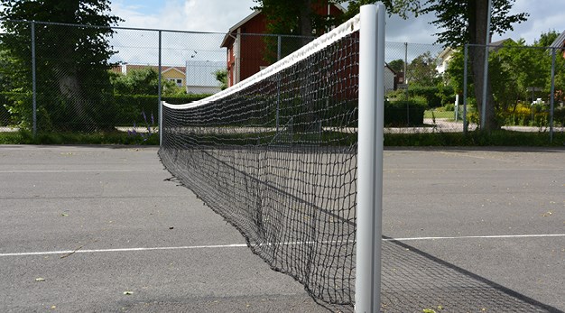Fotografiet är en närbild över ett tennisnät. Bilden är tagen från sidan så nätets ställning är i fokus. Marken är asfalt och i bakgrunden syns ett högt gallerstaket och en husknut. 