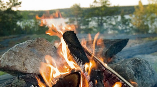 Vybild med en härlig lägereld i förgrunden och skog och vatten i bakgrunden.