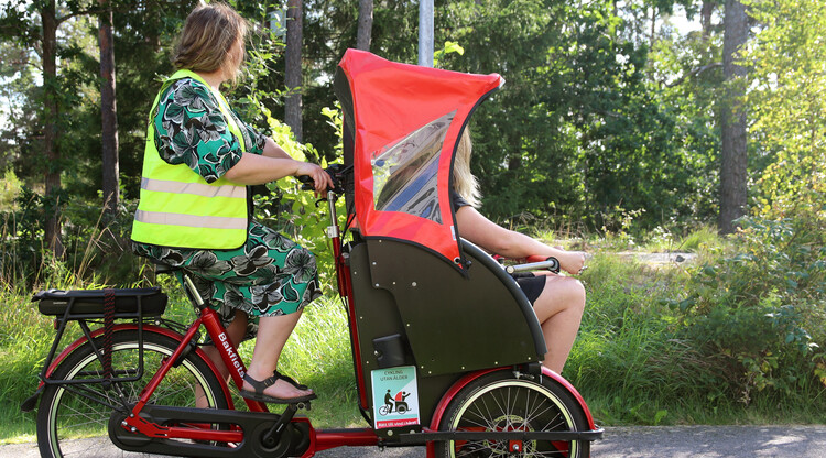 Kvinna i reflexväst cyklar med en brukare fram i en cykelvagn.
