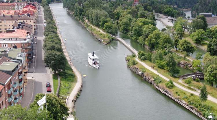 Foto över Göta kanal genom Trollhättan, en vit båt i vattnet och mycket träd och grönska runt om.