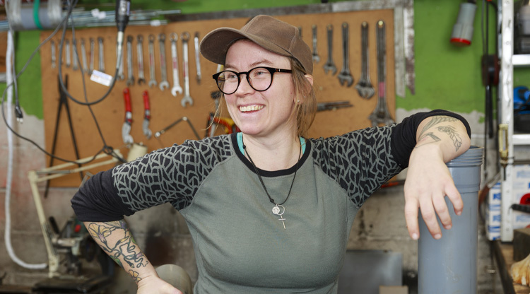 En leende person med keps och glasögon står i en verkstadsmiljö. Personen vilar ena armen mot ett rör och i bakgrunden syns en vägg med upphängda verktyg som skiftnycklar.