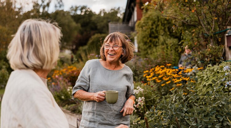 Två äldre kvinnor dricker kaffe och skrattar tillsammans i en trädgård. Foto.