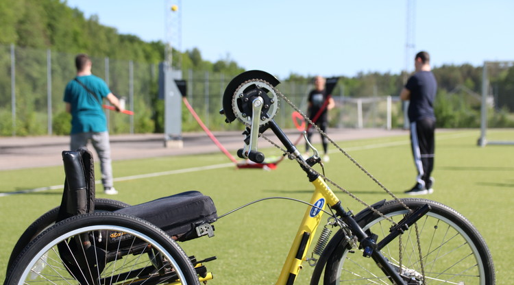 Handikappanpassad cykel man använder händerna för att föra cykeln framåt. I bakgrund skymtas personer spela badminton.