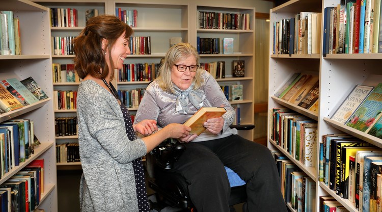 Bibliotekarie visar bok för besökare. I bakgrunden syns bokhyllor med böcker.