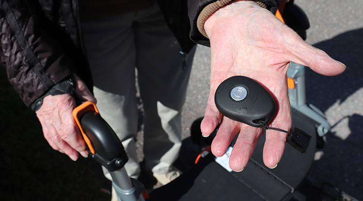 En äldre hand som håller ett mobilt trygghetslarm. I bakgrunden ser du en rollator.