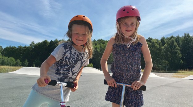 Fotografiet föreställer två flickor som ler in mot kameran. De står med varsin kickbike på Skateparken i Trollhättan. Tjejen till vänster är klädd i en tröja med en tiger på och gröna byxor. Hon har en orange hjälm. Tjejen till höger är klädd i en blå och rosablommig byxdress. Hon har en rosa hjälp. I bakgrunden syns hopp för kickbike och utkanten på en skog. 