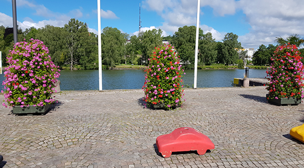 Fotografiet är taget utmed kanalen. I bild syns tre jättestora krukor med röda och lila blommor. I förgrunden syns en miniatyr SAAB som fungerar som en betongsugga.