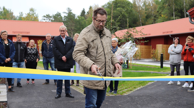 En hyresgäst klipper ett band utomhus under en invigningsceremoni på Stenlidenvägen. Bakom honom står flera besökare utspridda på en utegård och tittar på. I bakgrunden skymtas ett skogsparti och två radhuslängder.