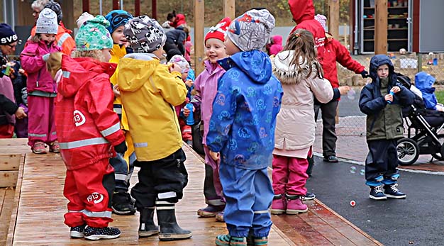Bilden föreställer ett flertal barn som står på en skolgård. Alla barnen har regnkläder i olika färger på sig. I förgrunden syns några barn som står i en ring och i bakgrunden syns lekande barn. 