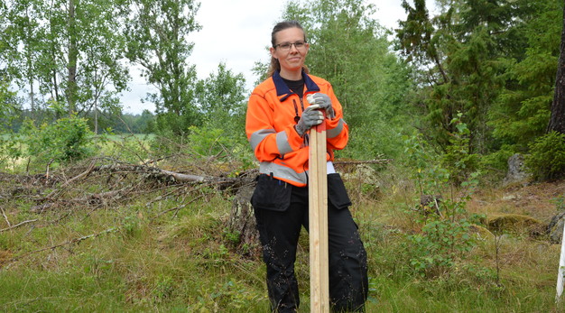 Bilden är ett fotografi på förrättningslantmätare Marianne. Marianne står mitt i bild omringad av skog och ler stort. Hon är iklädd en orange reflexjacka och svarta byxor. 