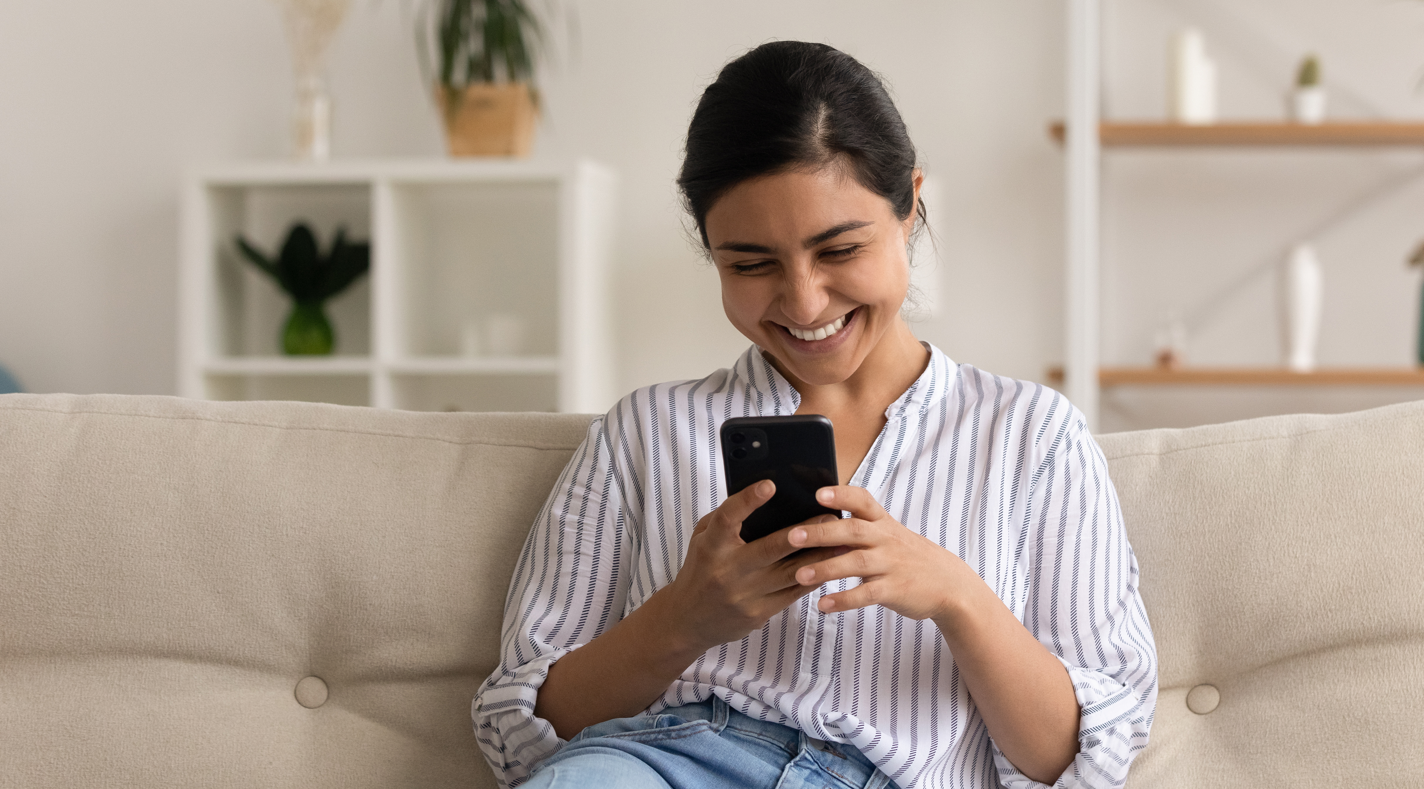 Ung kvinna sitter i en soffa i ett vardagsrum, tittar på telefonen och skrattar åt innehållet på skärmen.