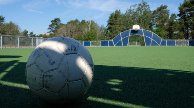 Bilden föreställer en fotbollsplan. I förgrunden till vänster är en fotboll i fokus. I bakgrunden syns en fotbollsplan med mål och även en basketkorg