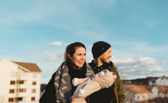 Två föräldrar med en nyfödd bebis utomhus med bostadsmiljö i bakgrunden
