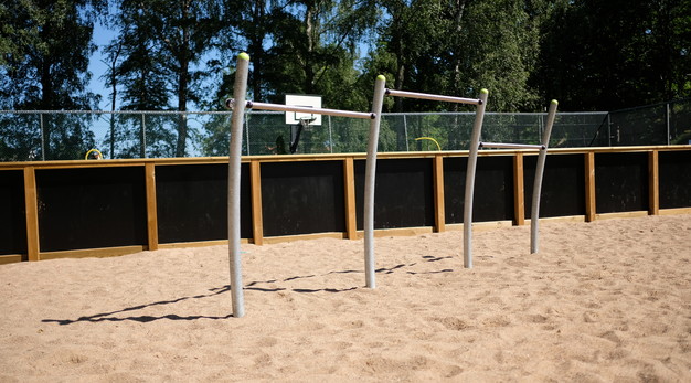 Bilden föreställer ett volträcke. Räcket är i stål med gula detaljer. Marken under är sand och i bakgrunden syns ett svart staket och en basketkorg i vitt med svarta detaljer. 