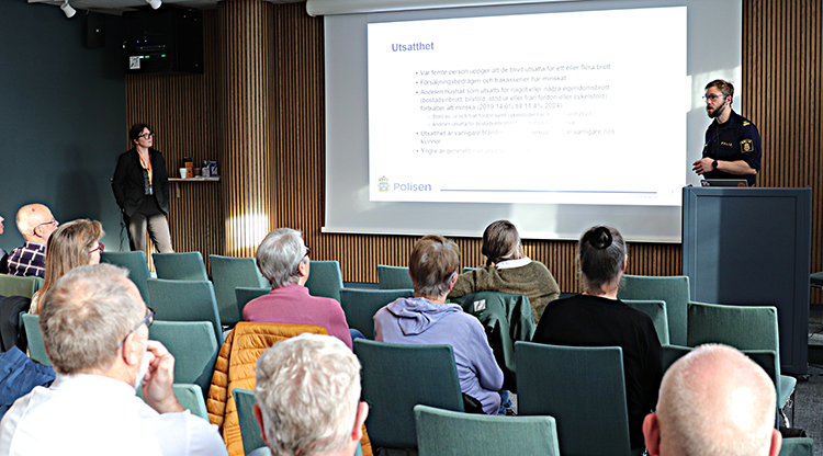 Polis i uniform står vid talarstol och visar bildspel på en stor skärm. Publik sitter framför honom. 