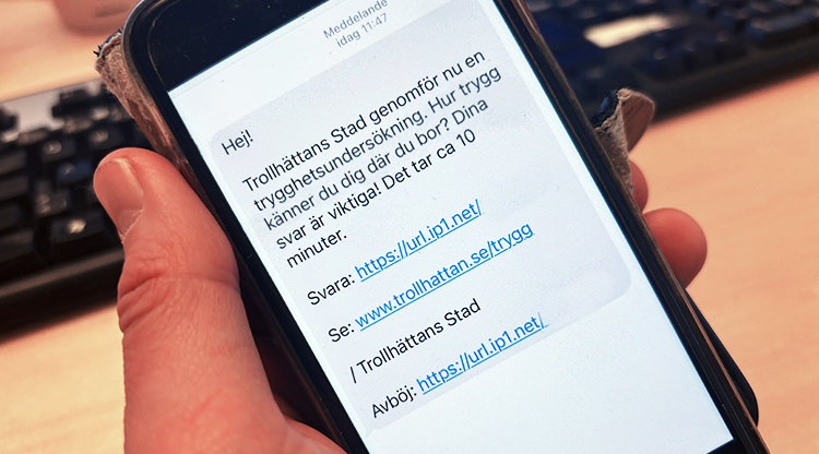 en hand håller en mobiltelefon. Man kan läsa ett sms med texten "Trollhättans Stad genomför nu en trygghetsundersökning."