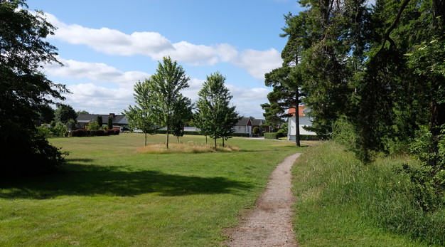 Bilden föreställer en liten grusstig som löper bredvid en park. Till vänster i bild är det en stor öppen yta med tre träd och till höger är det buskar och träd.
