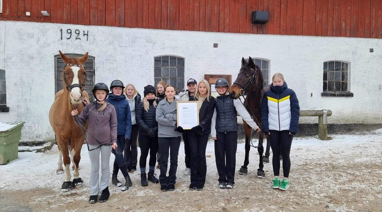 Ungdomar står utomhus vid stall med två hästar och håller upp ett diplom.