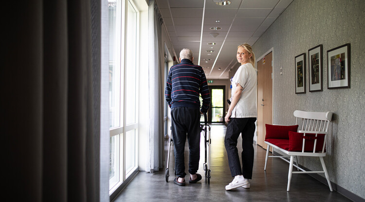 Kvinna i sjukhuskläder i korridor med man med rollator