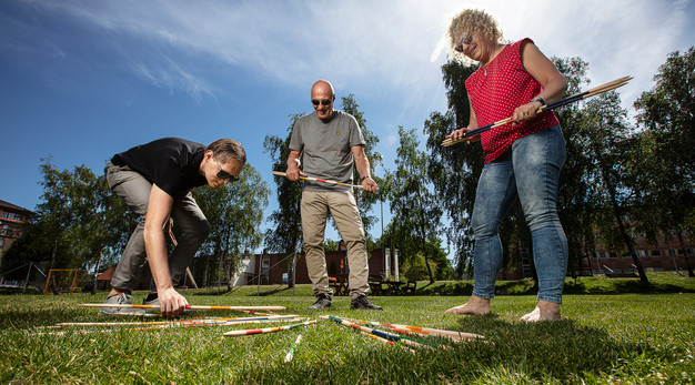 Bilden föreställer tre personer i en park som spelar plockepinn. Till vänster i bild står en man iklädd svart t-shirts och gråa byxor och böjer sig ned för att ta tag i en pinne. I mitten står en man i grå t-shirt och beiga byxor. Till höger i bild står en kvinna med röd blus och blåa jeans. Samtliga personer ler och de ser ut att ha roligt. 