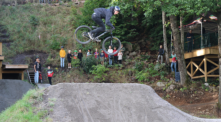 En man hoppar med en cykel i ett grushopp i skogen. M&aring;nga barn och unga tittar p&aring;.
