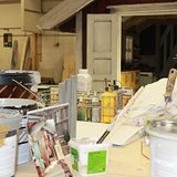 Fotografiet är en bild över ett skrivbord. Bordet är fullt med färgburkar, pendlar, inspirationsbilder och papper. I bakgrunden syns en stor kastrull och ingången till ett lekhus som ska placeras ut i Skrotnisses lekplats. 