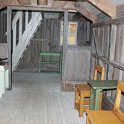 Foto är en bild över insidan på Nissestugan på Skrotnisses lekplats. Till vänster i bild syns ett kök med en spis. Spisen är vit och skåpsluckorna är gula. Till höger i bild syns två stolar och ett bord som står längst med väggen. Längst in i stugan syns en trappa som leder till övervåningen. Trappan är grå och under den står en lite hylla i grönt. 