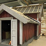 Bilden föreställer en lekstuga. Lekstugan består av två huskroppar. Huset är rött och har vita knutar. Huskroppen som är närmast i bild har en ingång med vita dubbeldörrar. Huskroppen närmast i bild sitter samman med den större huskroppen som har två plan. Huset håller på att byggas och just nu läggs det tak. 
