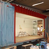Bilden föreställer byggandet av den scen som kommer placeras på Skrotnisses lekplats. Scenen byggs med hjälp av en gammal fraktcontainer. På containerns långsida har man sågat ut en stor öppning men behållit tak och väggarna på kortsidorna. Inuti containern står det en lång stege med en färgburk och penslar på.  